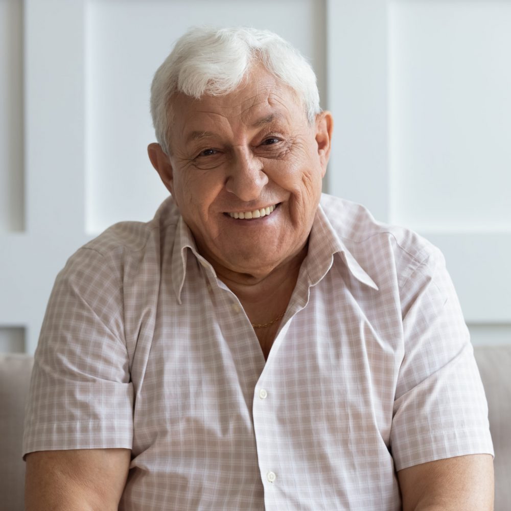 Close up headshot portrait of smiling mature man sit on couch at home look at camera posing for picture, happy positive senior male or optimistic grandfather feel good relax on comfortable sofa